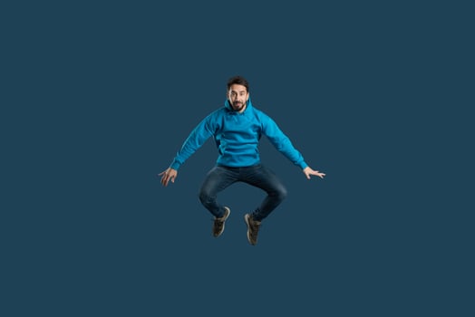 A man in a blue hoodie is mid-air, leaping with joy. The dark background emphasizes his vibrant outfit and energetic pose, showcasing an expression of excitement and freedom. photo