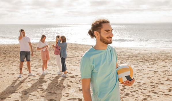 A young man stands on the beach, holding a volleyball, while friends engage in various activities nearby, enjoying a sunny day and the calming ocean waves. photo