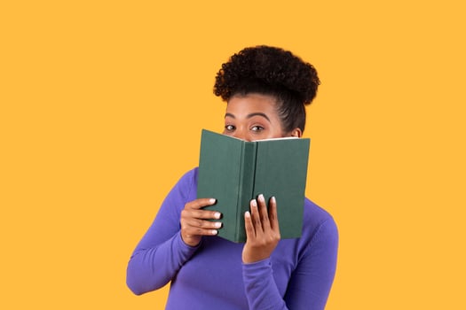 A young woman with curly hair holds a green book close to her face, peeking over it with a playful expression. The bright yellow background adds a cheerful vibe to the moment. photo