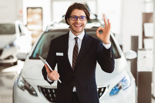 Car Sales. Smiling Handsome Salesman In Glasses Gesturing Okay Standing In Luxury Auto Store. Free Space photo