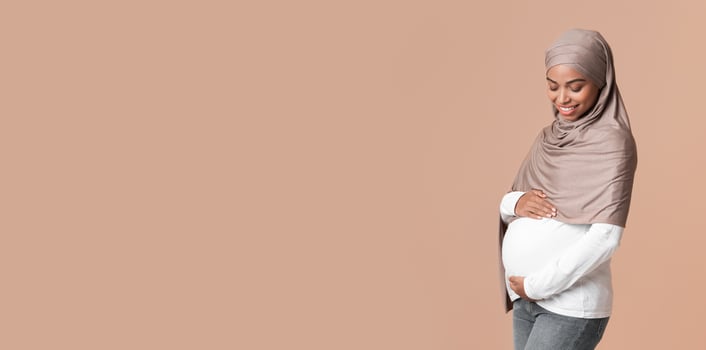 Happy black muslim woman in hijab expecting baby, tenderly touching her pregnant belly while posing over yellow background in studio, free space photo