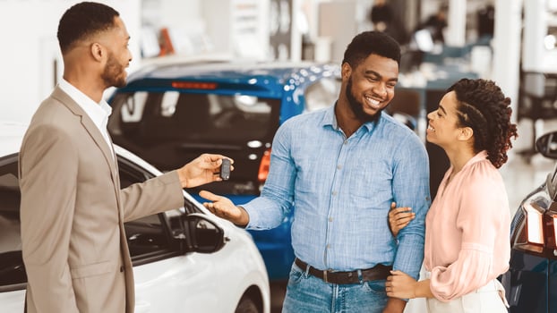 New Car. Salesman Giving Automobile Key To Happy Afro Couple Customers Standing In Auto Dealership Center. Panorama photo