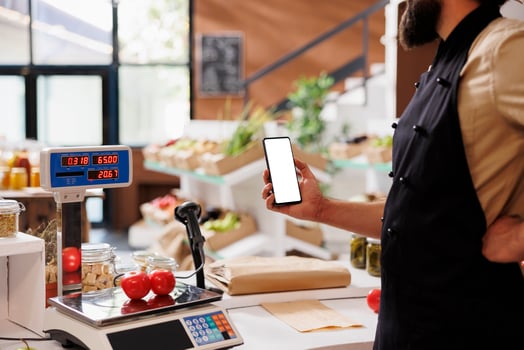 Storekeeper weighing fresh produce while gripping cellphone with blank mockup template at eco-friendly grocery store. Caucasian man wearing an apron and holding smartphone with white screen. photo