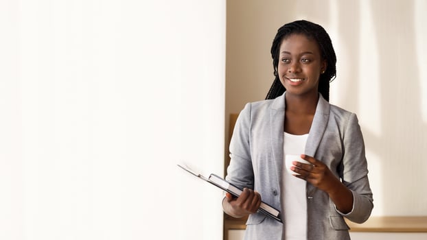 Pensive african secretary having coffee break near window in office, standing with clipboard, notebook and cup in hands, free space photo