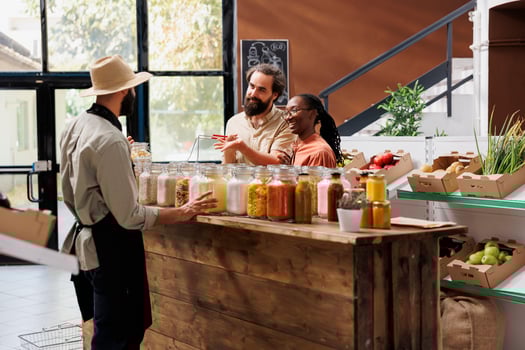 Male vendor offering advice to clients who are shopping for sustainable merchandise at eco friendly store. Owner of local supermarket describing food items in jars to the young multiracial couple. photo
