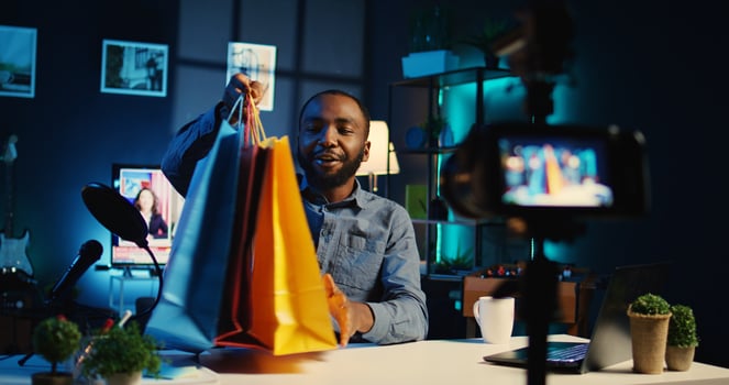 BIPOC content creator holding shopping bags, filming haul in personal apartment studio. Internet star unpacking clothing items he bought, reviewing fashion stores merchandise, handheld camera photo