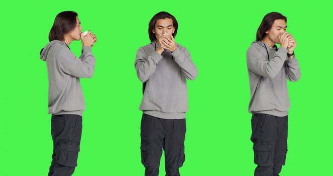 Person drinking cup of coffee on camera while standing in front of greenscreen template, enjoying caffeine refreshment. Young asian man holding hot beverage and feeling pleased. photo
