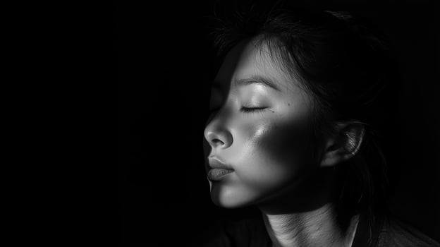 A close-up of a woman with closed eyes, illuminated by soft light casting shadows on her face in a dark environment photo