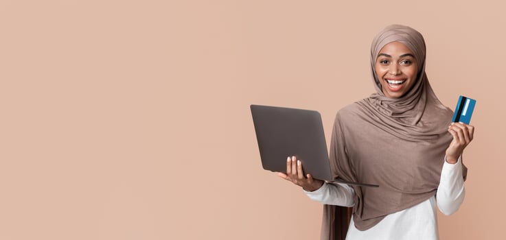 Online shopping concept. Joyful black muslim girl in hijab holding laptop and credit card and looking at camera, yellow background with free space photo