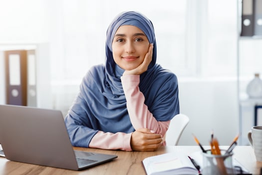 Muslim female entrepreneur sitting at desk in modern office and posing to camera, resting at workplace, free space photo