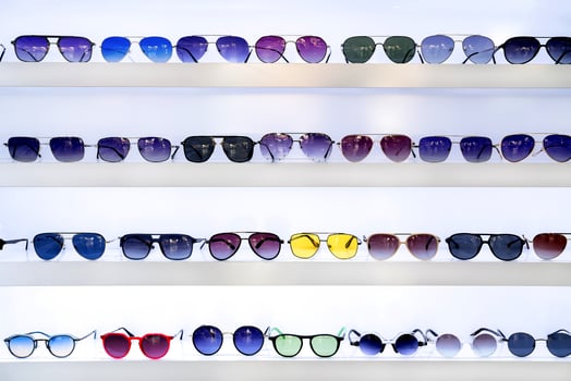 Rows of sunglasses in various styles and colors on white shelves, showcasing eyewear options, vibrant lens hues, fashion accessories, retail store presentation, organized selection, shopping ambiance photo