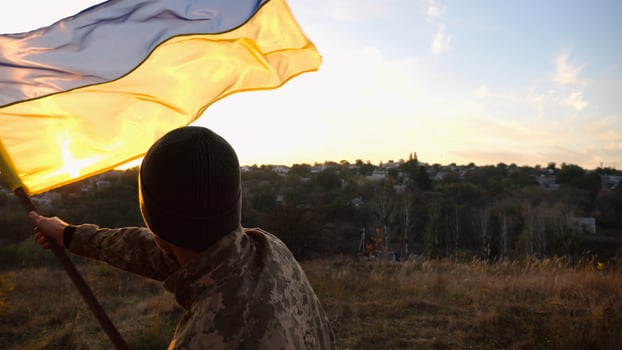 Young man in military uniform waving flag of Ukraine against beautiful sunset at background. Male ukrainian army soldier lifted national banner at countryside. Victory against russian aggression photo