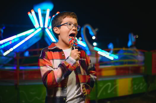 A cute caucasian boy eating a colorful lollipop standing against the background of a carousel in the evening at an amusement park or circus photo