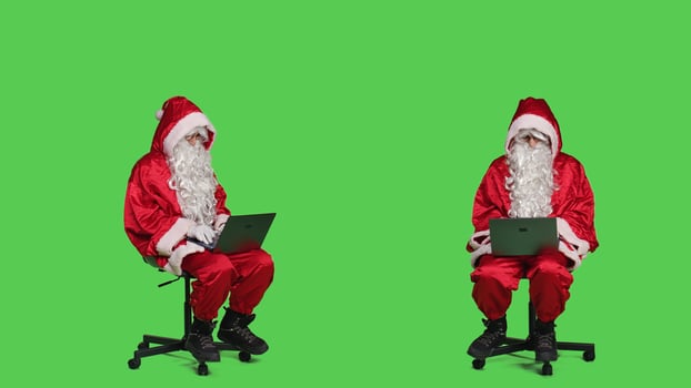 Cool saint nick using laptop on chair, sitting against full body greenscreen backdrop in studio. Father christmas working with modern portable pc, browsing online website in red costume. photo