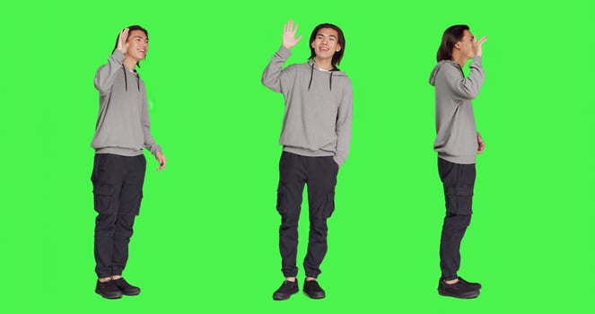 Asian man waving hello at people, greeting someone with a smile and confidence against full body greenscreen template. Young adult saying hi with a wave, smiling and feeling positive in studio. photo