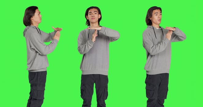 Person does timeout gesture with hands, asking for a break or pause against greenscreen in studio. Asian man showing disapproval and negative emotions on camera, stop gesture. photo