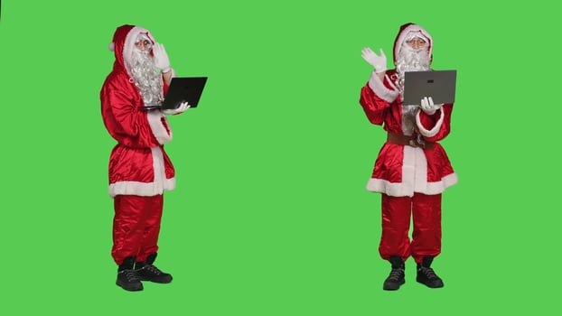 Saint nick waving at videocall on pc standing over full body greenscreen background, portraying december main character during christmas. Santa talks to people on online videocall. photo