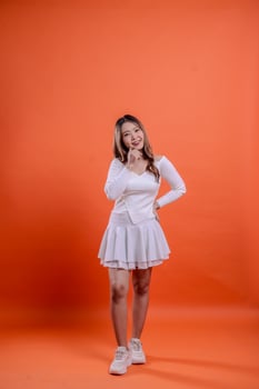 Full length beautiful Asian female in white top standing cheerful hands holding waist and chin isolated orange background. for the concept of fashion, beauty, advertising, photo