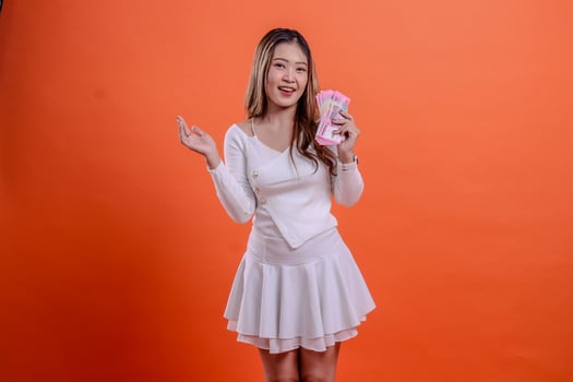 gesture of beautiful young asian woman in white top standing cheerfully holding rupiah cash and sideways isolated orange background. for business concept, competition, finance, reward photo