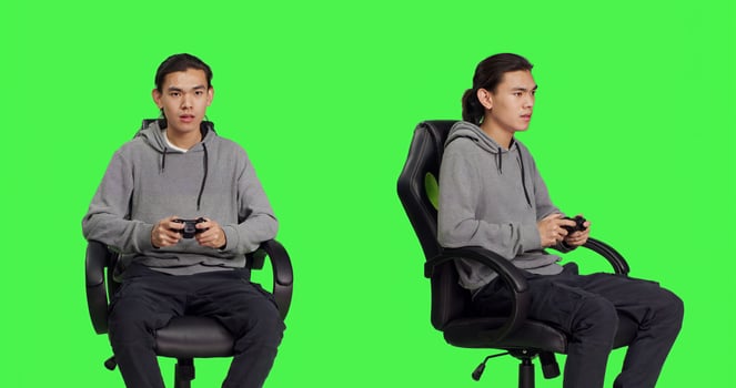 Asian guy using controller for gameplay, sitting on chair against greenscreen backdrop in studio. Young man enjoying online gaming plan with friends, gamer playing rpg games. photo
