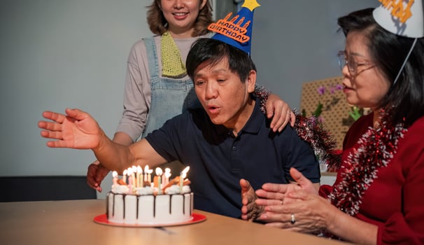 A man is blowing out candles on a birthday cake while two women watch photo