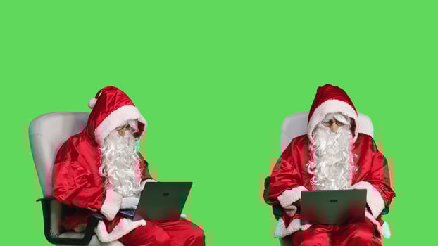 Trendy father christmas on laptop browsing website, sitting on chair with white and red iconic costume. Santa claus representing december festive holiday with long fake beard, use pc. photo