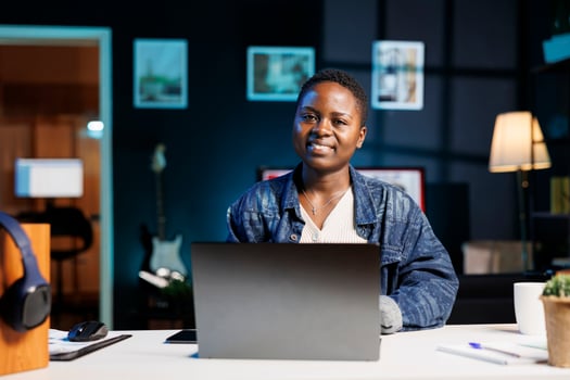 Portrait shot of smiling black woman working from home by using her personal computer. African american entrepreneur looking at camera and utilizing a digital laptop for research. photo