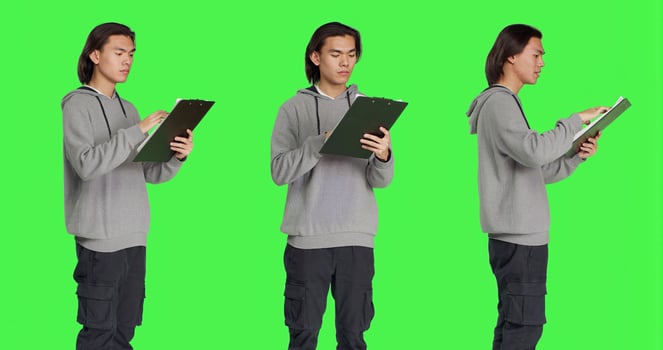 Asian guy reads information on files, thinking about annotations on clipboard papers or taking notes for completion. Young man writing on forms, standing against greenscreen backdrop. photo