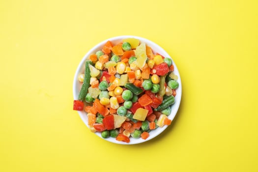 Chopped frozen vegetables in a plate on a yellow background. Corn peas pepper carrots. copy space. photo