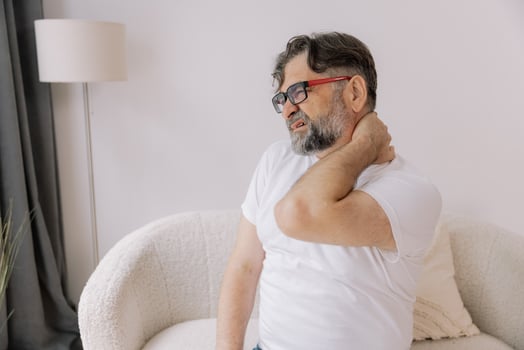 A senior man is sitting indoors, visibly suffering from neck pain. He is holding his neck with a pained expression, indicating discomfort. photo