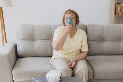 Sick senior woman has a viral infection or flu, labored breathing, asthma. An elderly female is using inhaler, nebulizer for self treatment sitting alone on the sofa at home photo