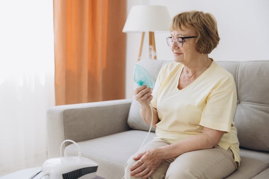Sick senior woman has a viral infection or flu, labored breathing, asthma. An elderly female is using inhaler, nebulizer for self treatment sitting alone on the sofa at home photo