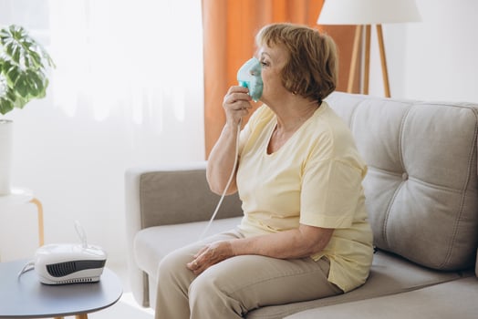 Sick senior woman has a viral infection or flu, labored breathing, asthma. An elderly female is using inhaler, nebulizer for self treatment sitting alone on the sofa at home photo