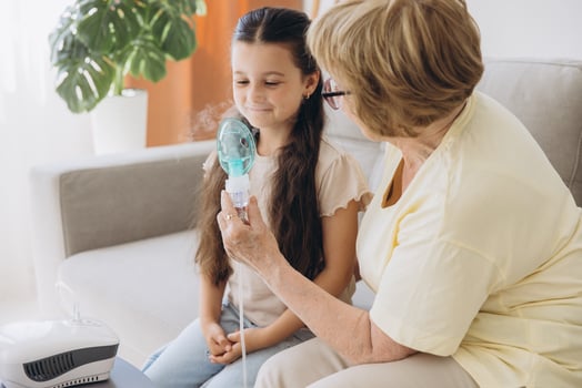 Little girl making inhalation with nebulizer with granny. Woman makes inhalation to a sick child while embracing her. Grandmother helping granddaughter with cold and flu to inahale nebuliser aerosol. photo