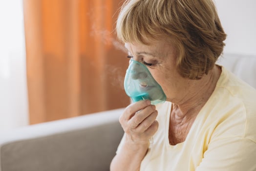 Sick senior woman has a viral infection or flu, labored breathing, asthma. An elderly female is using inhaler, nebulizer for self treatment sitting alone on the sofa at home photo