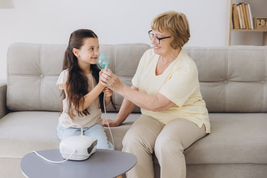 Little girl making inhalation with nebulizer with granny. Woman makes inhalation to a sick child while embracing her. Grandmother helping granddaughter with cold and flu to inahale nebuliser aerosol. photo