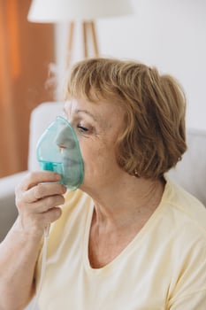 Sick senior woman has a viral infection or flu, labored breathing, asthma. An elderly female is using inhaler, nebulizer for self treatment sitting alone on the sofa at home photo