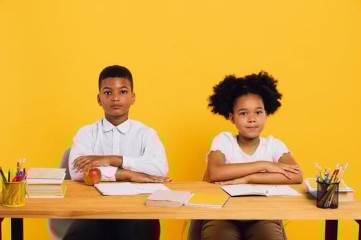 Happy african american schoolgirl and schoolboy sitting together at desk and studying on yellow background. Back to school concept. photo