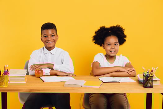Happy african american schoolgirl and schoolboy sitting together at desk and studying on yellow background. Back to school concept. photo