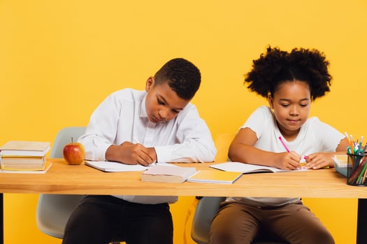 Happy african american schoolgirl and schoolboy sitting together at desk and studying on yellow background. Back to school concept photo