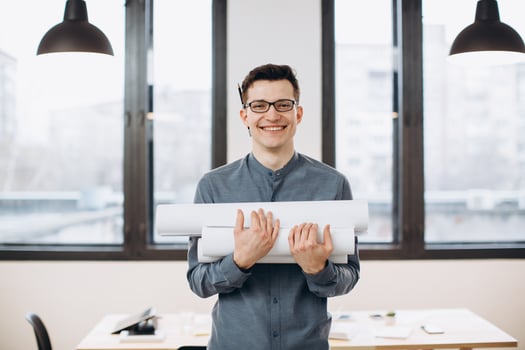 Attractive young man architect in glasses standing isolated over loft office background, carrying drawings, holding house scheme photo