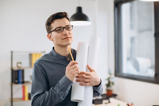 Attractive young man architect in glasses standing isolated over loft office background, carrying drawings, holding house scheme photo
