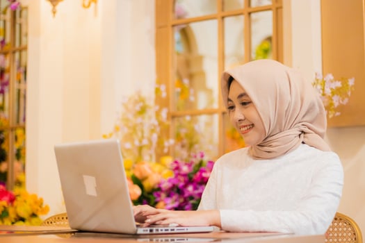 freelance beautiful young woman hijab wearing white muslim dress smiling typing message to her friend in front of laptop in luxury interior. for fashion, promotion, lifestyle, corporate content photo