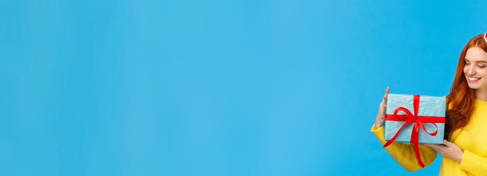 Holidays, love and family concept. Carefree tender and pretty redhead female holding cute wrapped box with gift, looking at present delighted, smiling happy, standing blue background joyful photo