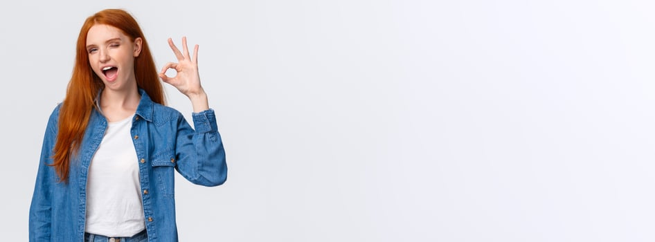 Waist-up portrait cheeky, confident and relaxed, chill redhead woman saying all alright, encourage everything okay, showing ok gesture wink and tongue, standing white background photo