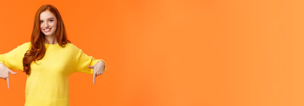 You need see this, check it out. Cheerful gorgeous redhead girl in yellow sweater, smiling and pointing down, recommend product, advertising shopping holidays sales, orange background photo
