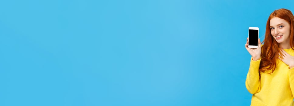 Impressed and fascinated redhead sassy woman touch heart and flirty gazing camera showing sexy guy photo she met dating app, holding smartphone with display facing camera, blue background