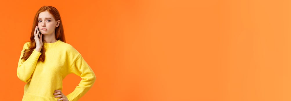 Girl catching anxiety calling someone. Perplexed or nervous cute redhead woman biting lip and looking away as having conversation on mobile phone, standing orange background sad photo