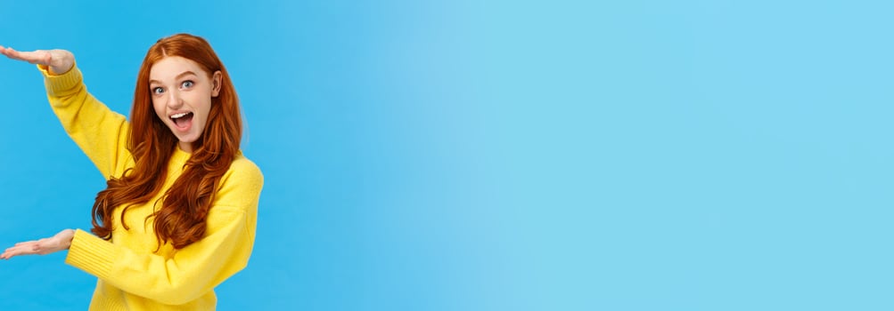 Excited lively good-looking redhead ecstatic girl smiling fascinated and amused, showing big, large object, shaping size with raised hands over blue background, standing joyful, impressed photo