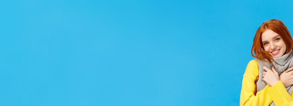 Winter holidays, sales and shopping concept. Waist-up portrait cute and lovely redhead woman wrap neck with soft warm scarf, tilt head smiling happily, embrace herself, standing blue background photo